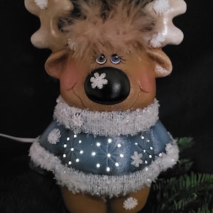 Puede incluir: Una figurita de reno de cer&aacute;mica con un su&eacute;ter azul y detalles de copo de nieve blanco. El reno tiene un cuerpo marr&oacute;n, cuernos blancos y una nariz negra. El su&eacute;ter est&aacute; decorado con copos de nieve blancos y tiene un borde de piel blanca. El reno tiene un copo de nieve blanco en la nariz y ojos azules.