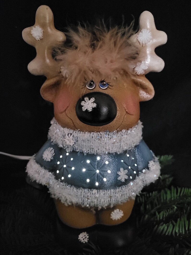 Puede incluir: Una figurita de reno de cer&aacute;mica con un su&eacute;ter azul y detalles de copo de nieve blanco. El reno tiene un cuerpo marr&oacute;n, cuernos blancos y una nariz negra. El su&eacute;ter est&aacute; decorado con copos de nieve blancos y tiene un borde de piel blanca. El reno tiene un copo de nieve blanco en la nariz y ojos azules.