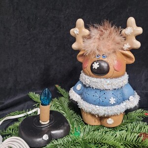 Puede incluir: Una figurita de reno de cer&aacute;mica con ojos azules, una cabeza peluda marr&oacute;n y un su&eacute;ter azul con copos de nieve blancos. El reno sostiene una bombilla con una bombilla de vidrio azul. El reno est&aacute; sentado en una rama de pino verde.