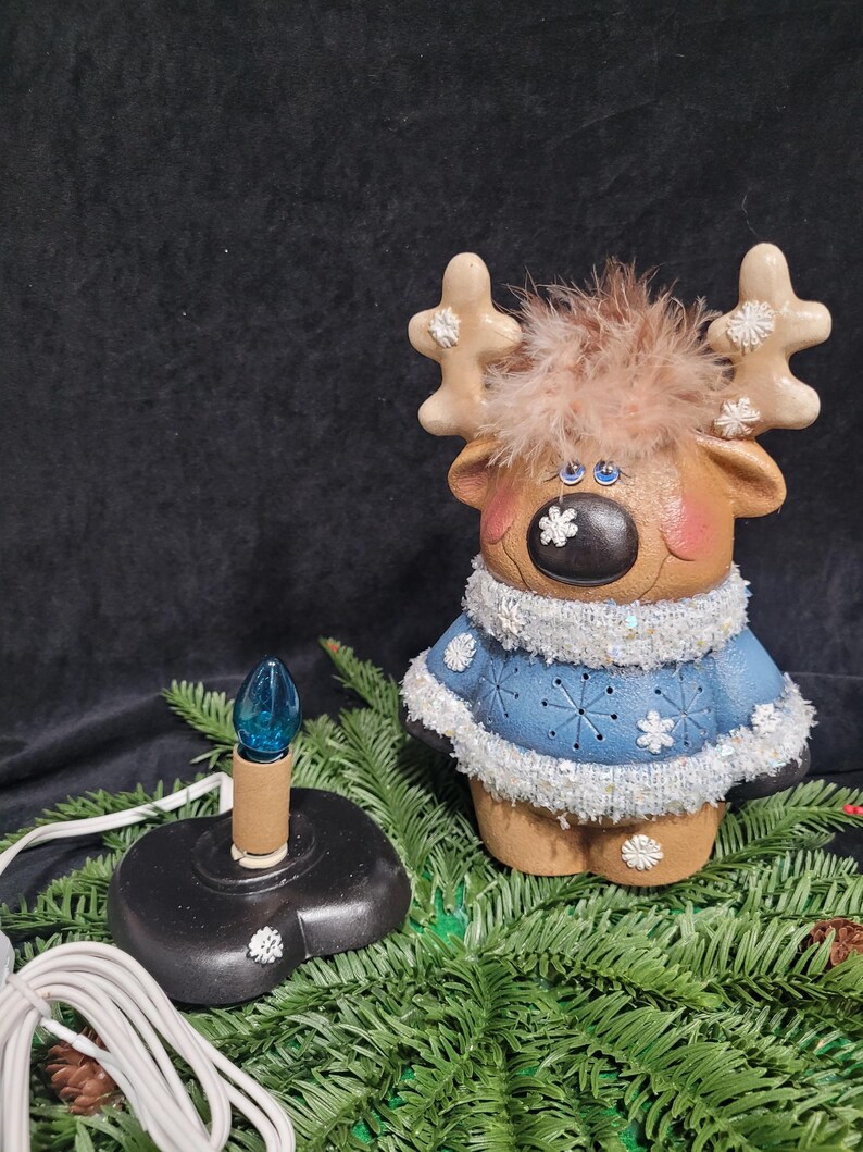 Puede incluir: Una figurita de reno de cer&aacute;mica con ojos azules, una cabeza peluda marr&oacute;n y un su&eacute;ter azul con copos de nieve blancos. El reno sostiene una bombilla con una bombilla de vidrio azul. El reno est&aacute; sentado en una rama de pino verde.