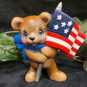 K&ouml;nnte beinhalten: Eine braune Teddyb&auml;r-Figur, die eine kleine amerikanische Flagge mit blauen Sternen und roten und wei&szlig;en Streifen h&auml;lt. Der B&auml;r tr&auml;gt eine blaue Glitzer Schleife.