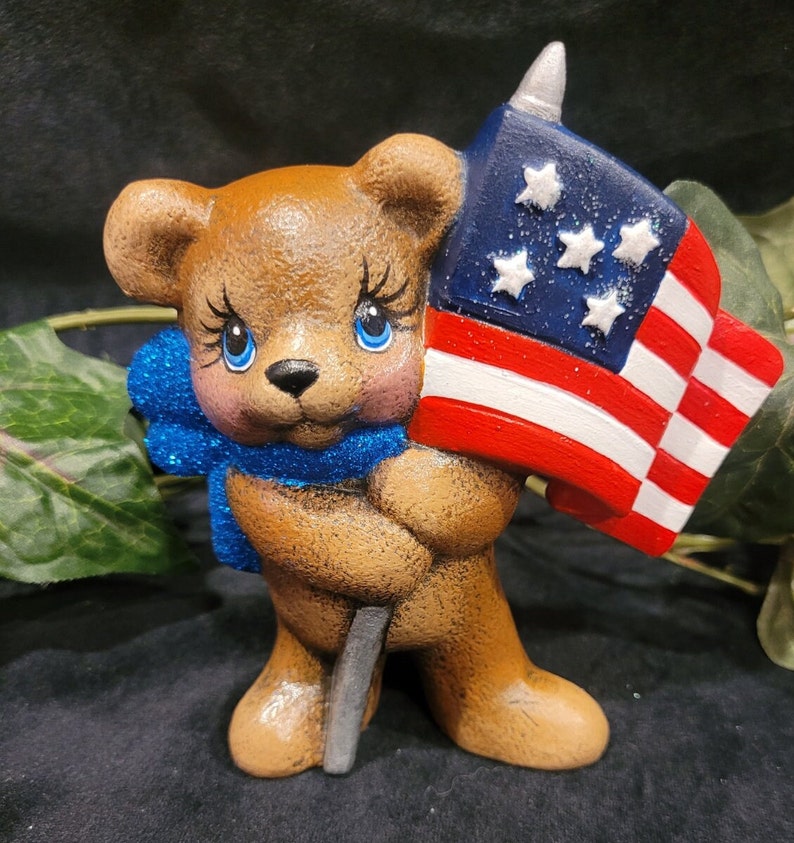 K&ouml;nnte beinhalten: Eine braune Teddyb&auml;r-Figur, die eine kleine amerikanische Flagge mit blauen Sternen und roten und wei&szlig;en Streifen h&auml;lt. Der B&auml;r tr&auml;gt eine blaue Glitzer Schleife.