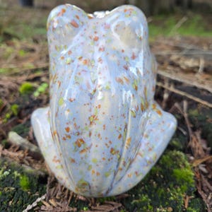Puede incluir: Una figurita de rana de cer&aacute;mica con un esmalte azul claro y un patr&oacute;n moteado de naranja, amarillo y blanco.