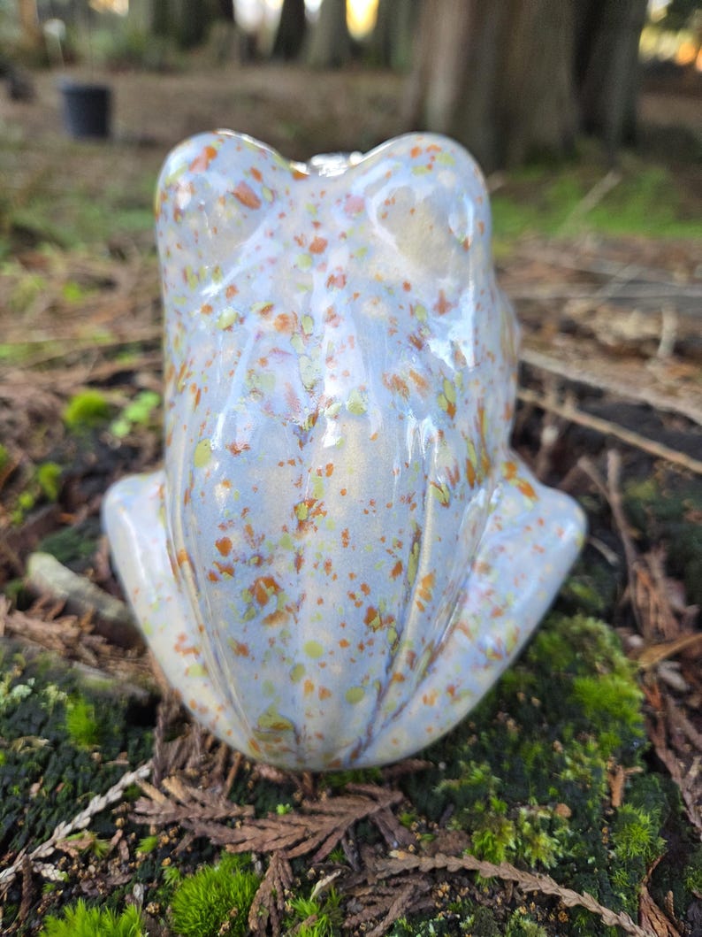 Puede incluir: Una figurita de rana de cer&aacute;mica con un esmalte azul claro y un patr&oacute;n moteado de naranja, amarillo y blanco.