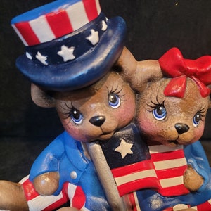 May include: Two ceramic teddy bears dressed in red, white, and blue patriotic outfits. The bear on the left is wearing a blue and white hat with stars and holding a small American flag. The bear on the right is wearing a red bow and holding a larger American flag.