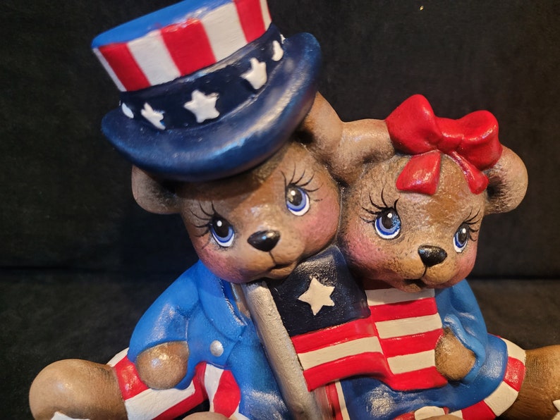 May include: Two ceramic teddy bears dressed in red, white, and blue patriotic outfits. The bear on the left is wearing a blue and white hat with stars and holding a small American flag. The bear on the right is wearing a red bow and holding a larger American flag.
