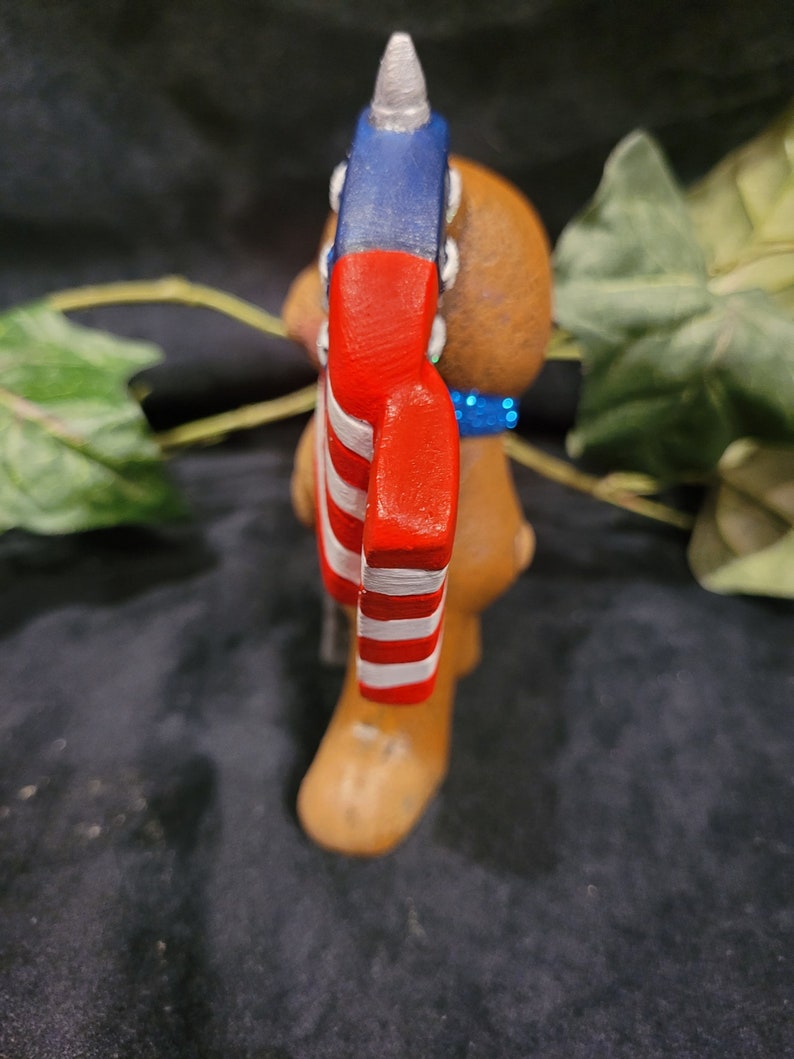 K&ouml;nnte beinhalten: Eine braune Keramikfigur eines B&auml;ren, der einen roten, wei&szlig;en und blauen Hut mit einer spitzen Spitze tr&auml;gt. Der Hut ist mit Sternen und Streifen verziert, die an die amerikanische Flagge erinnern.