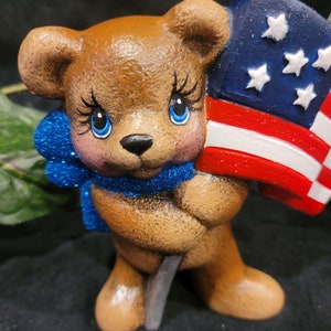 K&ouml;nnte beinhalten: Eine braune Teddyb&auml;r-Figur, die eine kleine amerikanische Flagge h&auml;lt. Der B&auml;r hat blaue Augen und eine blaue Glitzer Schleife um den Hals.