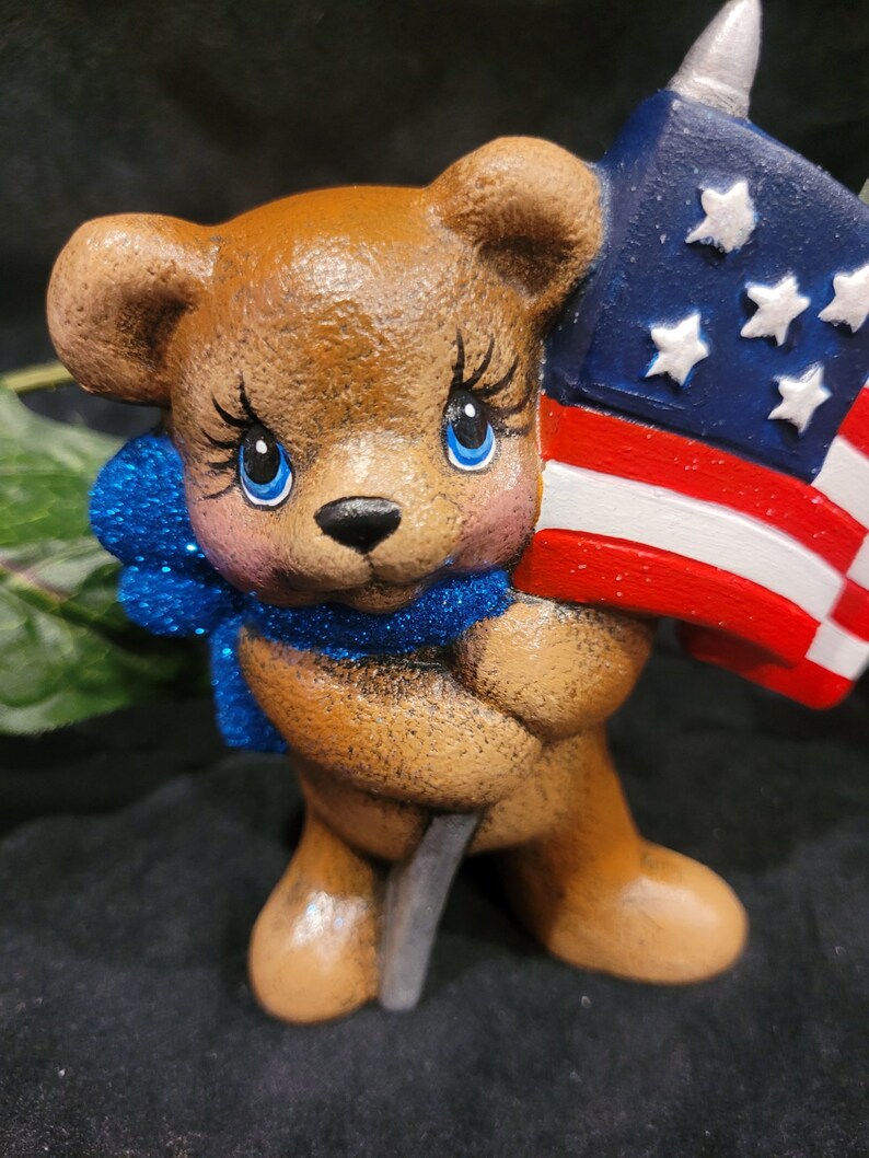 K&ouml;nnte beinhalten: Eine braune Teddyb&auml;r-Figur, die eine kleine amerikanische Flagge h&auml;lt. Der B&auml;r hat blaue Augen und eine blaue Glitzer Schleife um den Hals.
