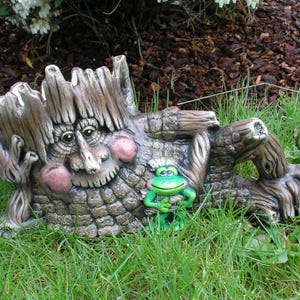 May include: A decorative garden sculpture shaped like a reclining tree spirit with a textured bark-like surface, a face with pink cheeks, and a small bright green frog figure resting against its side, all rendered in shades of brown and gray.