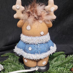 Puede incluir: Una figurita de reno de cer&aacute;mica con un su&eacute;ter azul y blanco, cuernos marrones y una pluma marr&oacute;n en la cabeza. El reno est&aacute; iluminado con un cable blanco.