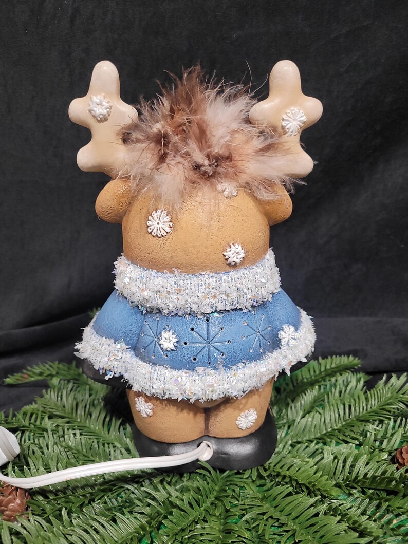 Puede incluir: Una figurita de reno de cer&aacute;mica con un su&eacute;ter azul y blanco, cuernos marrones y una pluma marr&oacute;n en la cabeza. El reno est&aacute; iluminado con un cable blanco.