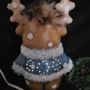 Puede incluir: Una figurita de reno de cer&aacute;mica con un su&eacute;ter azul y ribetes blancos. El reno tiene pelaje marr&oacute;n y cuernos blancos. El su&eacute;ter tiene un patr&oacute;n de copo de nieve y est&aacute; iluminado con luces blancas.