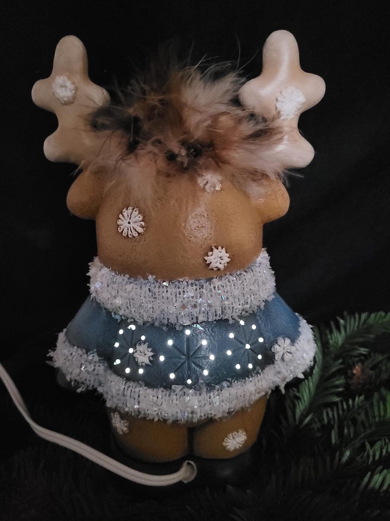 Puede incluir: Una figurita de reno de cer&aacute;mica con un su&eacute;ter azul y ribetes blancos. El reno tiene pelaje marr&oacute;n y cuernos blancos. El su&eacute;ter tiene un patr&oacute;n de copo de nieve y est&aacute; iluminado con luces blancas.