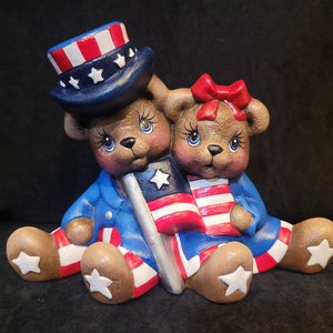 May include: Two ceramic teddy bears dressed in red, white, and blue patriotic outfits. The bear on the left is wearing a blue hat with red and white stripes and white stars. Both bears are holding American flags.