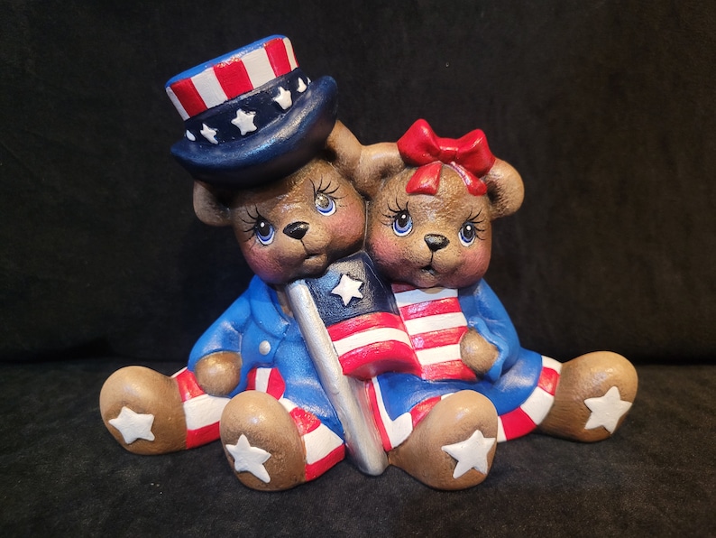 May include: Two ceramic teddy bears dressed in red, white, and blue patriotic outfits. The bear on the left is wearing a blue hat with red and white stripes and white stars. Both bears are holding American flags.