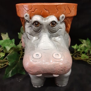 May include: A ceramic planter shaped like a hippopotamus with a brown pot on top. The hippopotamus has a gray body with pink cheeks and brown eyes.