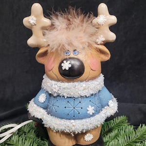 Puede incluir: Una figurita de reno de cer&aacute;mica con un su&eacute;ter azul, ribete de piel blanca y una cabeza de plumas marrones. El reno tiene una nariz negra, ojos azules y detalles de copos de nieve blancos.