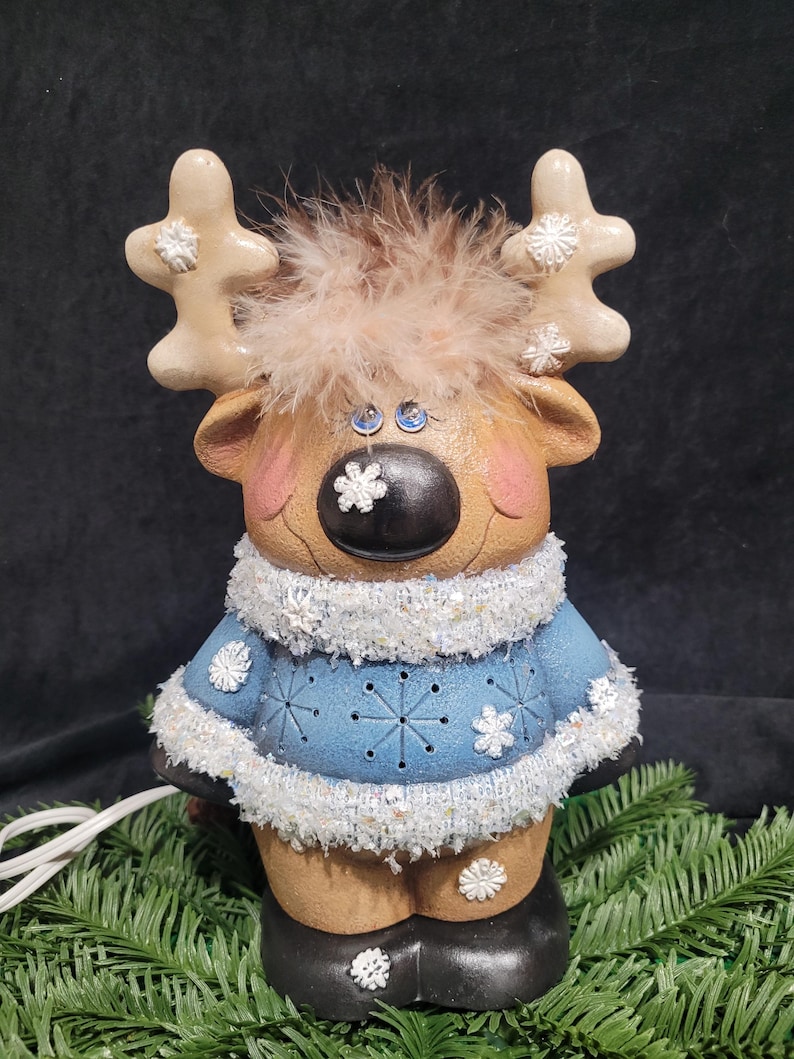 Puede incluir: Una figurita de reno de cer&aacute;mica con un su&eacute;ter azul, ribete de piel blanca y una cabeza de plumas marrones. El reno tiene una nariz negra, ojos azules y detalles de copos de nieve blancos.