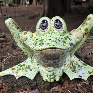 May include: A ceramic frog figurine with a light green glaze and dark green and blue speckles. The frog has large, prominent eyes with dark pupils, raised front limbs, and wide, flat feet. The surface has a glossy finish and a smooth texture.