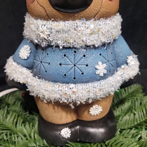 Puede incluir: Una figurita de reno de cer&aacute;mica que lleva un su&eacute;ter azul con copos de nieve blancos y un ribete de piel blanca. El reno tiene una cara marr&oacute;n con una nariz negra y copos de nieve blancos en las mejillas.