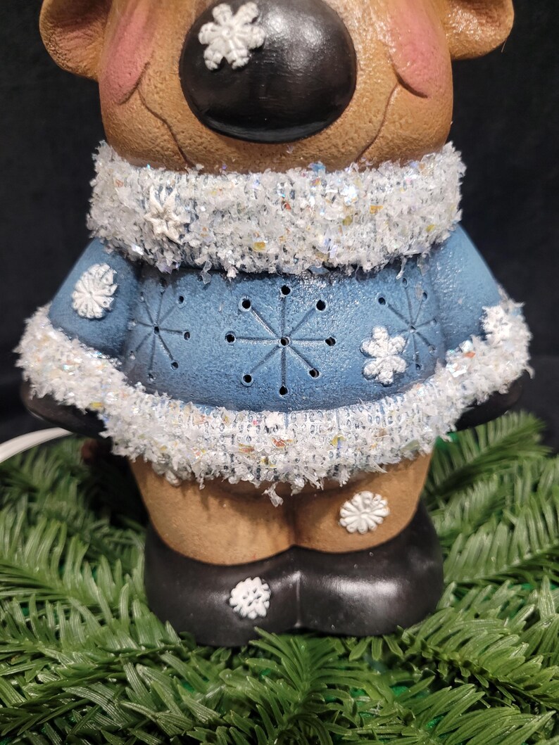 Puede incluir: Una figurita de reno de cer&aacute;mica que lleva un su&eacute;ter azul con copos de nieve blancos y un ribete de piel blanca. El reno tiene una cara marr&oacute;n con una nariz negra y copos de nieve blancos en las mejillas.