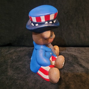 May include: A ceramic bear figurine wearing a blue, red, and white hat and matching outfit. The bear is sitting with its legs crossed and has a patriotic theme.