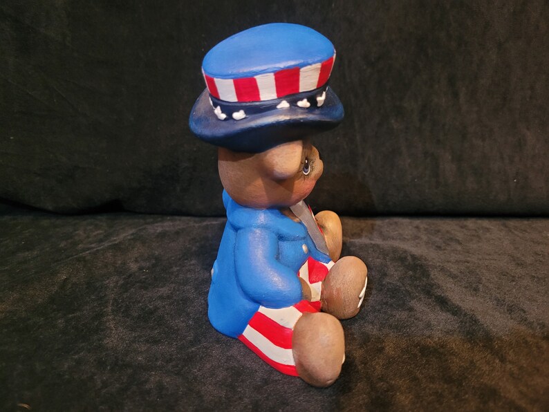 May include: A ceramic bear figurine wearing a blue, red, and white hat and matching outfit. The bear is sitting with its legs crossed and has a patriotic theme.