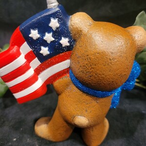 K&ouml;nnte beinhalten: Eine braune Keramik-B&auml;renfigur, die eine kleine amerikanische Flagge mit einer silbernen Spitze oben h&auml;lt. Der B&auml;r tr&auml;gt ein blaues, glitzerndes Band um den Hals.