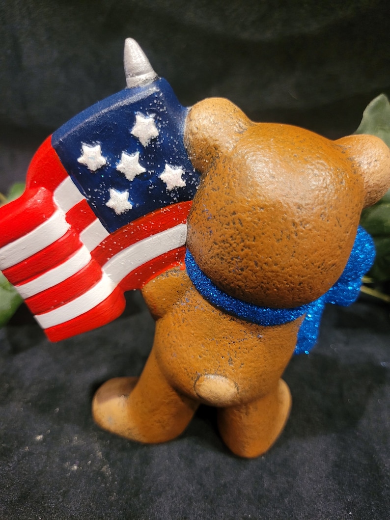 K&ouml;nnte beinhalten: Eine braune Keramik-B&auml;renfigur, die eine kleine amerikanische Flagge mit einer silbernen Spitze oben h&auml;lt. Der B&auml;r tr&auml;gt ein blaues, glitzerndes Band um den Hals.