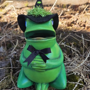 May include: A green ceramic frog figurine wearing a black felt witch hat with a small green accent on top. The frog has blue eyes, a wide mouth, and a black ribbon bow tied around its neck. The figure is seated with its hands folded together.