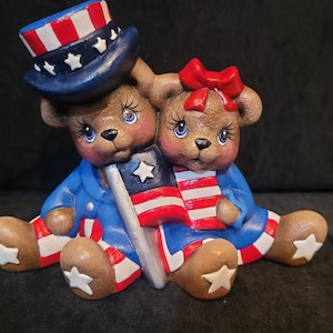 May include: Two ceramic teddy bears dressed in red, white, and blue patriotic outfits. The bear on the left is wearing a blue and white hat with stars and holding a small American flag. The bear on the right is wearing a red bow and holding a small American flag.