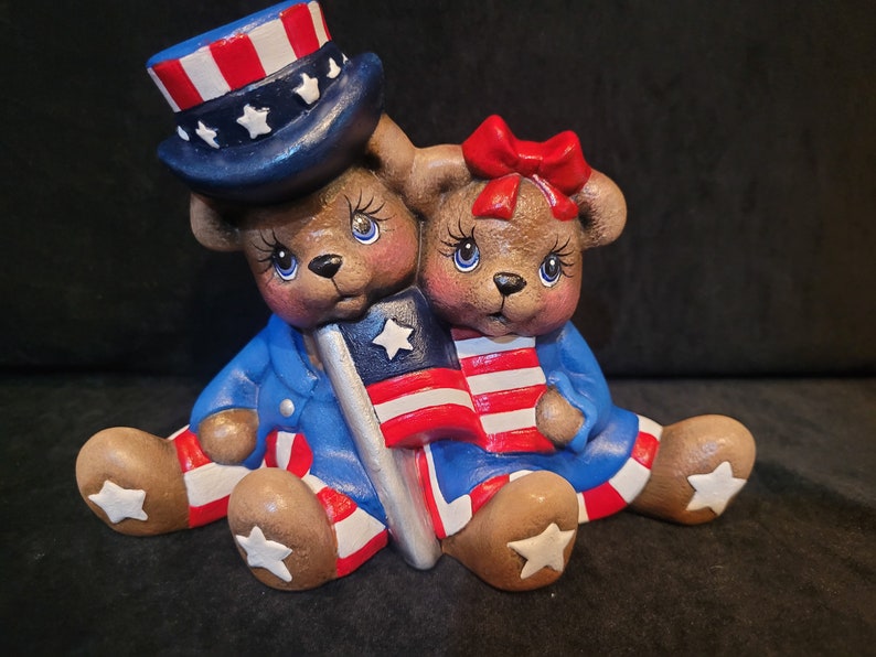 May include: Two ceramic teddy bears dressed in red, white, and blue patriotic outfits. The bear on the left is wearing a blue and white hat with stars and holding a small American flag. The bear on the right is wearing a red bow and holding a small American flag.