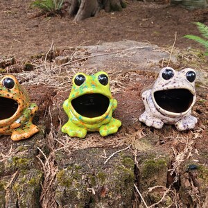 Puede incluir: Tres figuritas de rana de cer&aacute;mica con la boca abierta, cada una con un patr&oacute;n de color diferente: verde con manchas amarillas, naranja con manchas negras y blanco con manchas negras.