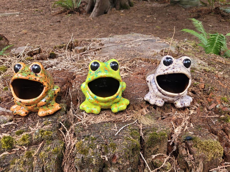 Puede incluir: Tres figuritas de rana de cer&aacute;mica con la boca abierta, cada una con un patr&oacute;n de color diferente: verde con manchas amarillas, naranja con manchas negras y blanco con manchas negras.