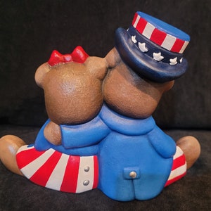 May include: Two brown teddy bears wearing red, white, and blue patriotic outfits. The bear on the right is wearing a blue hat with red and white stripes and white stars. The bear on the left is wearing a red bow and a red, white, and blue striped skirt.
