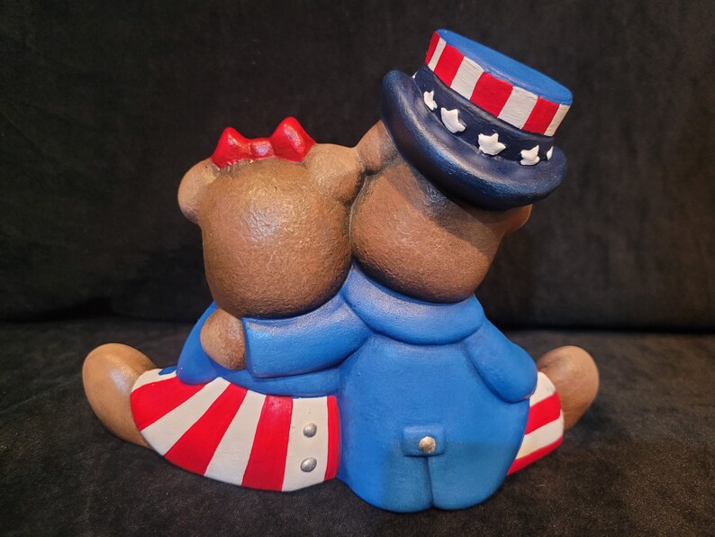 May include: Two brown teddy bears wearing red, white, and blue patriotic outfits. The bear on the right is wearing a blue hat with red and white stripes and white stars. The bear on the left is wearing a red bow and a red, white, and blue striped skirt.