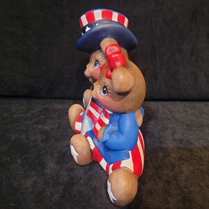 May include: Two brown teddy bears wearing red, white, and blue patriotic outfits. The bear on the left is wearing a blue and white striped hat with red stars. The bear on the right is wearing a blue jacket with red and white stripes.