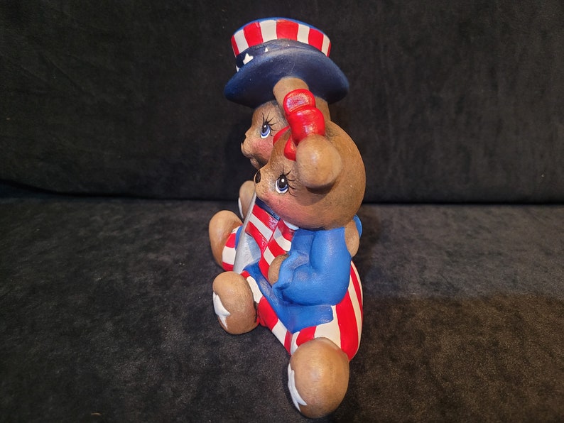 May include: Two brown teddy bears wearing red, white, and blue patriotic outfits. The bear on the left is wearing a blue and white striped hat with red stars. The bear on the right is wearing a blue jacket with red and white stripes.
