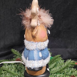 Puede incluir: Una figurita de reno de cer&aacute;mica con un su&eacute;ter azul y blanco, ribete de brillantina blanca y cuernos marrones con puntas blancas. El reno lleva un sombrero marr&oacute;n con un pomp&oacute;n blanco y tiene una cara marr&oacute;n claro con mejillas rosadas. La figurita est&aacute; iluminada con un cable blanco.