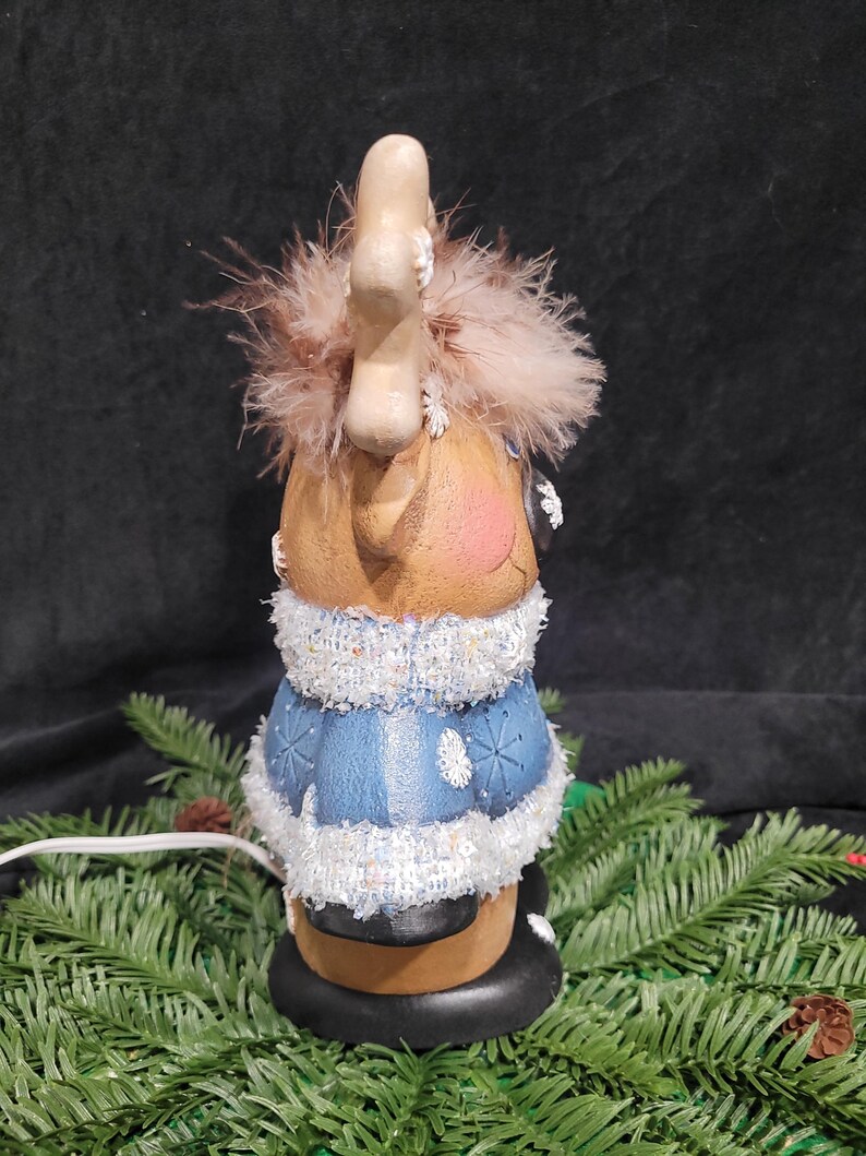 Puede incluir: Una figurita de reno de cer&aacute;mica con un su&eacute;ter azul y blanco, ribete de brillantina blanca y cuernos marrones con puntas blancas. El reno lleva un sombrero marr&oacute;n con un pomp&oacute;n blanco y tiene una cara marr&oacute;n claro con mejillas rosadas. La figurita est&aacute; iluminada con un cable blanco.