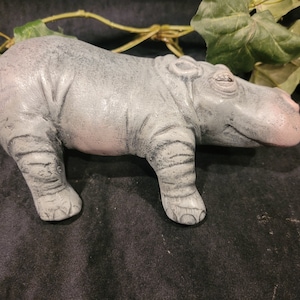 May include: A gray and pink ceramic hippopotamus figurine. The hippopotamus is lying down with its head turned to the side. The figurine has a detailed texture and a realistic appearance.