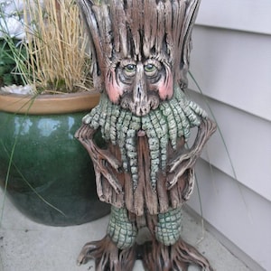 May include: A standing garden sculpture shaped like a tree spirit with a textured brown bark body, a long green mossy beard, and large green eyes. The figure has pointed ears, pink cheeks, and root-like feet, crafted with detailed wood grain textures.