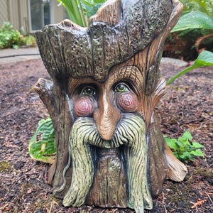 May include: A whimsical tree stump planter with a carved face, featuring a long white beard, a brown nose, and green eyes. The planter is made of a brown and green ceramic material and has a rustic, woodland aesthetic.