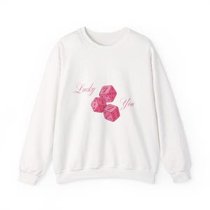 May include: A white crewneck sweatshirt featuring a graphic print of three pink dice with the number seven on each face. The text Lucky You is printed in a cursive pink font on either side of the dice, centered on the front of the garment.