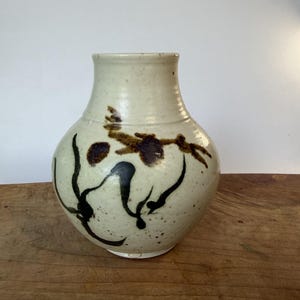 May include: A cream-colored ceramic vase with a speckled finish and a rounded body. The vase features hand-painted dark brown and black abstract floral or botanical brushstroke designs around the center and a narrow, smooth neck opening.