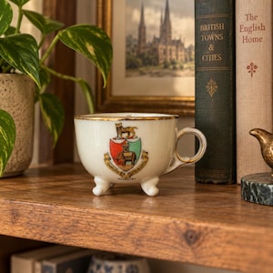 May include: A white ceramic teacup with a gold-trimmed rim and handle, featuring three small feet. The front displays a colorful heraldic crest with a tiger, a castle, and an elephant, accompanied by the text Camera Principis Coventry.