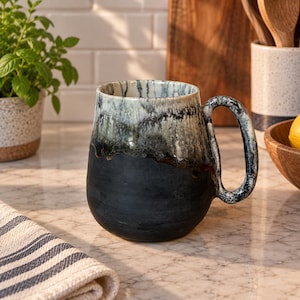 Peut inclure: Une tasse en céramique présentant un design bicolore avec une base noire mate et un vernis brillant de style goutte à goutte dans des tons de blanc, gris et noir sur la partie supérieure. La tasse a un corps arrondi et une anse marbrée assortie.