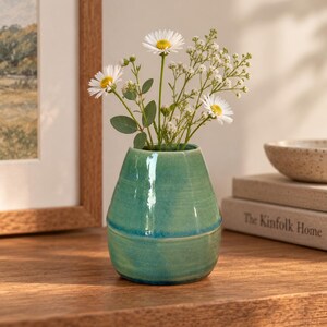 May include: A small, tapered ceramic vase with a glossy, variegated teal and green glaze. The vase features a subtle horizontal ridge around its midsection and contains a bouquet of white daisies and small white filler flowers with green stems.