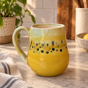 May include: A ceramic mug with a rounded body and a curved handle. The mug features a glossy finish with a gradient of pale green at the rim transitioning into a vibrant yellow base, accented by dark blue speckled dots around the upper section.